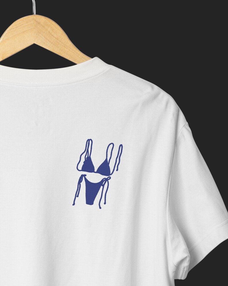 May include: White t-shirt with a navy blue bikini graphic on the back. The bikini design is a simple line drawing, showing the top and bottom with tie details. The shirt is hanging on a wooden hanger against a dark background.