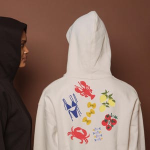 May include: Two hooded sweatshirts, one black and one off-white. The off-white hoodie has colourful illustrations of a lobster, bikini, pasta, lemons, tomatoes, crab, and fish. The background is a solid brown.