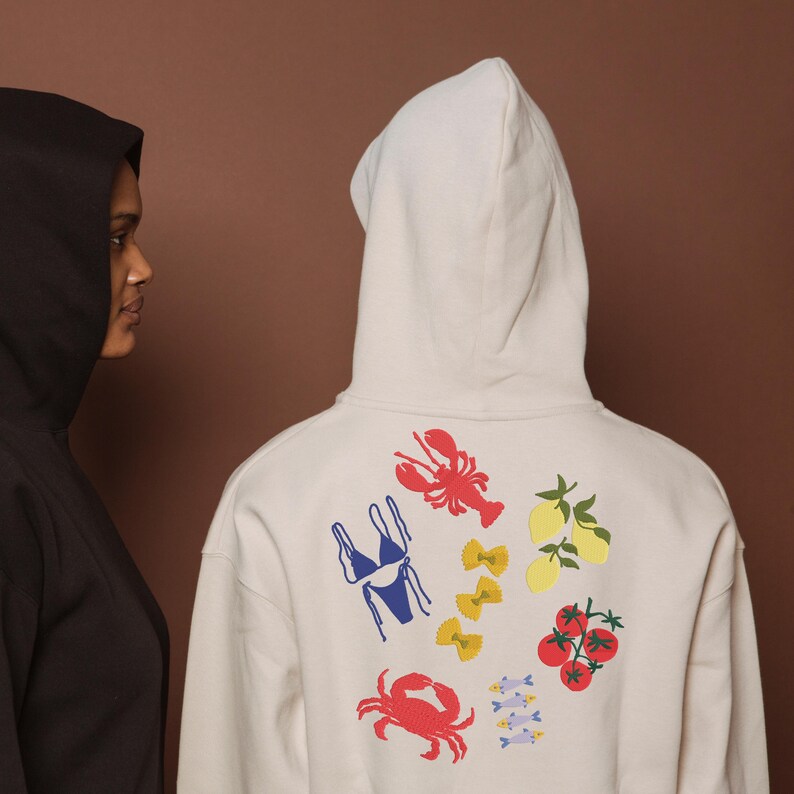 May include: Two hooded sweatshirts, one black and one off-white. The off-white hoodie has colourful illustrations of a lobster, bikini, pasta, lemons, tomatoes, crab, and fish. The background is a solid brown.