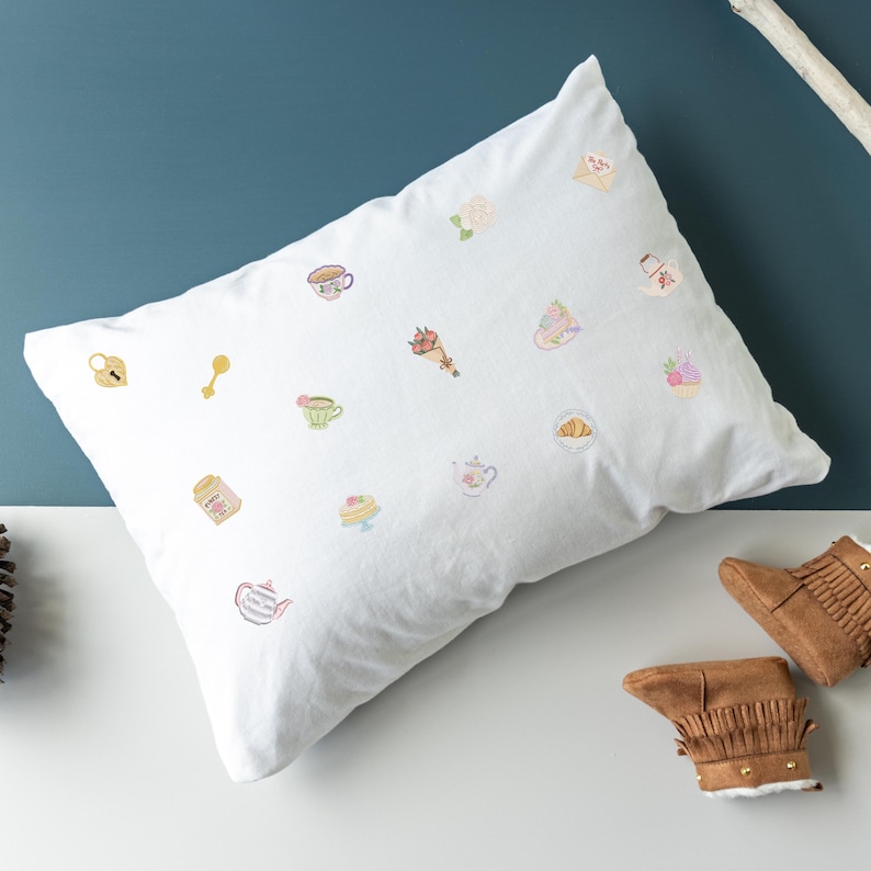 May include: White pillow with embroidered pastel-colored illustrations of teacups, teapots, pastries, and other charming objects. The pillow is on a white surface with a blue background. Brown fringed baby boots are in the bottom right corner.