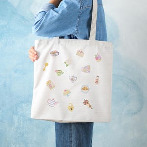 May include: Cream-colored tote bag featuring various pastel-colored illustrations of teacups, teapots, pastries, and floral elements. The bag is held by a person wearing a denim jacket, and the background is a light blue.