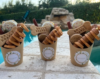 Könnte beinhalten: Drei braune Pappbecher gefüllt mit Leckerlis für Hunde. Die Becher haben einen weißen Kreis mit dem Text "Brit's Boards" darauf. Die Leckerlis sind in verschiedenen Formen und Größen, darunter Knochen, Ringe und Kekse.