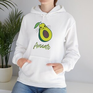 Pode incluir: Moletom branco com capuz com um design de abacate verde. O abacate tem um rosto de desenho animado fofo e a palavra "Avocado" está escrita em verde abaixo.