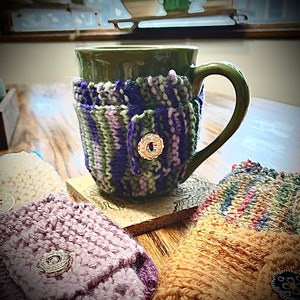May include: A green ceramic mug with a purple, green, and pink knitted cozy. The cozy has a button on the front. There are three other knitted cozies in the image.
