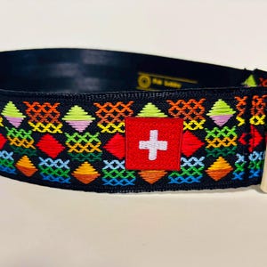 May include: Black dog collar with a vibrant embroidered pattern. The design includes diamonds and crosses in red, orange, yellow, green, purple, and blue. A red square with a white cross is sewn on the collar. It has a silver metal buckle.