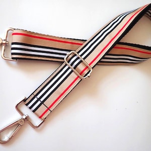 May include: A beige, black, and red striped fabric strap with silver metal clasps and hooks. The strap is designed to be used as a replacement strap for a bag.