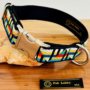 Könnte beinhalten: Ein schwarzes und mehrfarbiges Hundehalsband mit einer silbernen Schnalle und einem D-Ring. Das Halsband hat ein geometrisches Muster mit roten, gelben, grünen, blauen und weißen Streifen. Das Halsband wird von "Fob Lobby USA" hergestellt.
