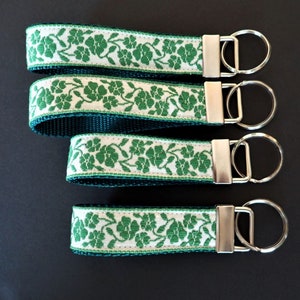Llavero con forma de TRÉBOL. Llavero con diseño de jacquard verde sobre blanco. Correa verde. Herrajes de níquel plateado. Pulsera. Ancho de 2,5 cm. Tamaño a elegir. Llavero irlandés con la inscripción San Patricio
