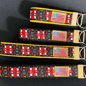 Op de afbeelding: Vier sleutelhangers met een zwart, rood, wit en blauw geometrisch patroon en een kleine Amerikaanse vlag patch. Elke sleutelhanger heeft een goudkleurige metalen sluiting en sleutelring. De sleutelhangers zijn in aflopende volgorde gerangschikt.