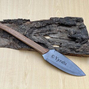 May include: A long, silver knife with a wooden handle. The blade is engraved with the text "© Yjcraftz". The knife is resting on a piece of weathered wood.