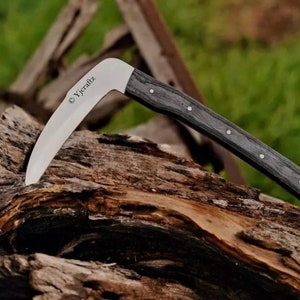 Puede incluir: Un cuchillo de hoja curva con un mango de madera gris oscuro. La hoja es plateada y tiene grabado el texto "Yjcraftz". El cuchillo está apoyado sobre un trozo de madera.