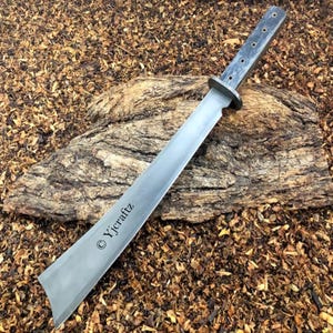 Puede incluir: Una espada larga de color plateado con un mango de madera negro. El mango tiene una serie de agujeros y la hoja es curva. La espada está apoyada en un trozo de madera con corteza marrón. El texto "©Yjcraftz" está grabado en la hoja.