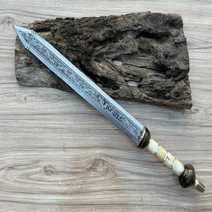 Puede incluir: Una espada plateada con un diseño intrincado en la hoja y un mango de hueso. La espada está grabada con el texto "Yeraftz".