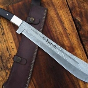 Puede incluir: Un cuchillo largo de color plateado con mango negro y funda de cuero. La hoja tiene un patrón de Damasco y es muy afilada. El cuchillo está sobre una superficie de madera.