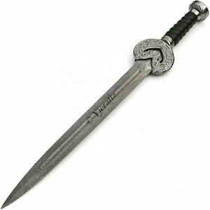 Puede incluir: Una espada plateada con una guarda decorativa en forma de corazón y un mango de cuero negro. La hoja es de acero damasco con un patrón ondulado.