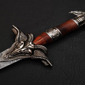 Puede incluir: Una espada decorativa con un mango de madera y una hoja plateada. La hoja está decorada con un diseño detallado de una cabeza de carnero. El mango está envuelto en cuero marrón y tiene una pomo de metal plateado. La espada está sobre un fondo negro.