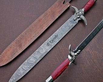 May include: A Damascus steel sword with a red wooden handle and a decorative horned design. The sword is accompanied by a brown leather sheath. The image also shows a smaller matching dagger. The text "Yjcraftz" is visible on the sword.