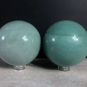May include: Two polished green aventurine spheres on clear acrylic stands. The spheres have a smooth, reflective surface and vary slightly in shade, one being lighter than the other. The background is dark, highlighting the gemstones.