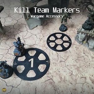 Könnte beinhalten: Zwei schwarze Plastik-Kill-Team-Marker mit einem sechszackigen Stern-Design. Die Marker sind mit 1 und 2 nummeriert. Die Marker befinden sich auf einer rissigen, beigefarbenen Oberfläche. Der Text "Kill Team Markers" und "Wargame Accessory" ist im Bild sichtbar.