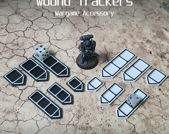 Könnte beinhalten: Ein Set mit 13 weißen und schwarzen Wundmarkern für Tabletop-Kriegsspiele. Die Marker sind in Form von kleinen Booten mit jeweils drei Fächern gestaltet. Ein weißer Würfel liegt auf einem der Marker.