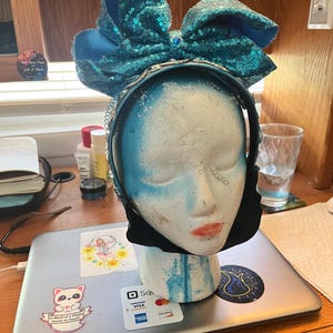 Puede incluir: Una diadema con lazo de lentejuelas turquesas sobre una cabeza de maniquí. La diadema tiene un lazo grande y está sujeta a una banda negra. La cabeza del maniquí está pintada de azul y blanco con labios rojos.
