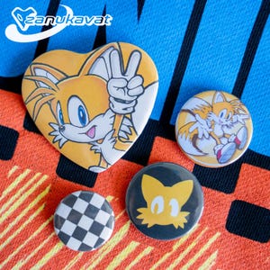 Può includere: Una collezione di quattro spille colorate con il personaggio Tails della serie Sonic the Hedgehog. Le spille includono un design a forma di cuore, un bottone rotondo con Tails in una posa dinamica, un bottone a scacchi in bianco e nero e un bottone con una silhouette gialla.