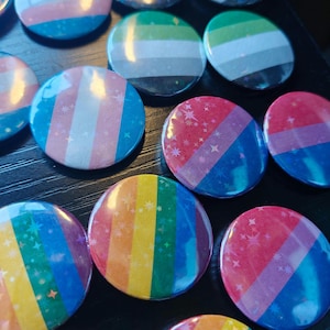 Könnte beinhalten: Eine Sammlung runder Ansteckbuttons mit verschiedenen Pride-Flaggen-Farbmustern. Jeder Button hat eine glänzende Oberfläche mit einem dezenten holografischen Sternenmuster. Die Designs zeigen horizontale Streifen in Regenbogenfarben.