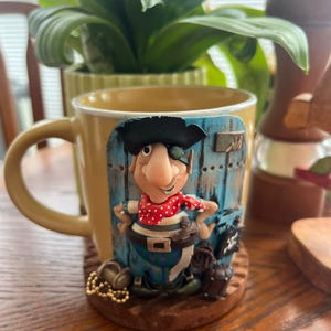 May include: A yellow ceramic mug with a pirate-themed design. The mug features a cartoon pirate with an eye patch, red polka-dot scarf, and black hat. The pirate is standing in front of a blue wooden door. A gold chain and treasure chest are at the base.