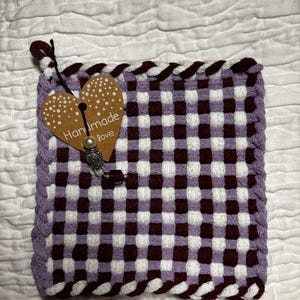 Op de afbeelding: Een vierkante geweven pannenlap met een ruitpatroon in tinten lavendel, wit en donker kastanjebruin. De randen zijn afgewerkt met een gevlochten boord. Een hartvormig bruin papieren label met de tekst Handmade love is met een touwtje bevestigd.