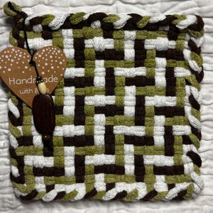 Op de afbeelding: Een vierkante geweven pannenlap met een geometrisch patroon in olijfgroen, donkerbruin en wit. Een hartvormig label met de tekst "Handmade with love" is bevestigd aan een zwart koord met een kleine crèmekleurige kraal en een donkerbruine houten kraal.