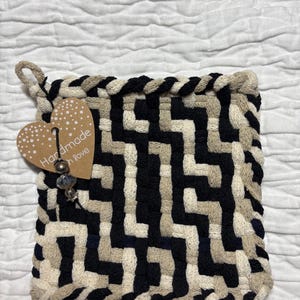 Op de afbeelding: Handgemaakte vierkante onderzetter met een geometrisch patroon in zwart, beige en crème. De randen zijn gevlochten. Een hartvormig label met "Handmade with Love" is bevestigd.