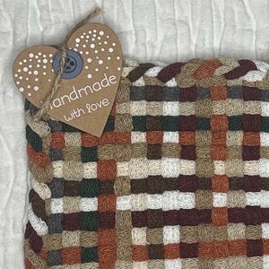 May include: Close-up of a woven coaster with a mix of brown, beige, green, and white squares. A heart-shaped tag with the words "handmade with love" is attached with a button and twine. The coaster is likely handmade.