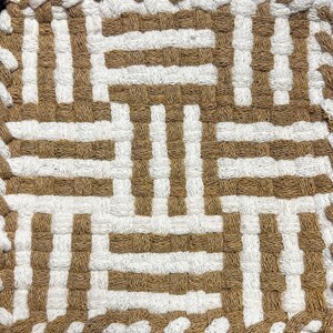 May include: A square textile coaster or mat featuring a woven pattern of alternating tan and white fabric strips. The design consists of a geometric grid with vertical and horizontal segments creating a repeating block structure across the surface.