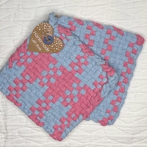 Op de afbeelding: Twee vierkante geweven pannenlappen met een geblokt patroon in lichtblauw en roze garen. Een hartvormig bruin papieren label is met een blauwe knoop aan de hoek bevestigd, met de witte gedrukte tekst, Handmade with love.