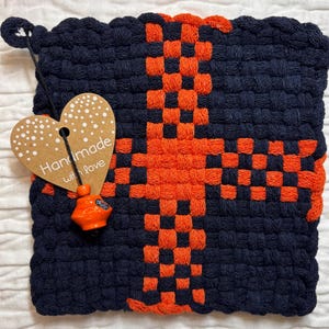 May include: A square, woven potholder in navy blue and orange. The potholder features a cross design in orange against a navy blue background. A heart-shaped tag with the words "Handmade with love" is attached to the potholder.