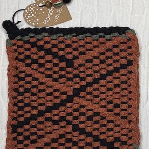 May include: Woven square pot holder in a checkerboard pattern of black and rust-orange. The edges are trimmed with black and a thin strip of green. A heart-shaped tag with the words "Handmade with love" is attached.