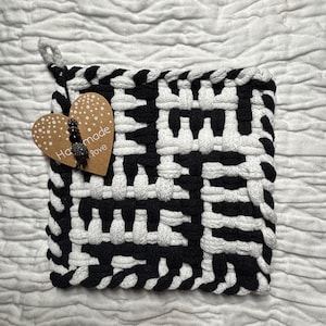 Op de afbeelding: Een vierkante, zwart-witte geweven pannenlap met een ophanglus. Een houten hartvormig label met de woorden "Handmade Love" is bevestigd. De pannenlap is gemaakt van dik, duurzaam materiaal.