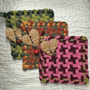 May include: Three woven square pot holders in shades of green, orange, and pink. Each pot holder has a different color scheme and a heart-shaped tag that says "Handmade with love".