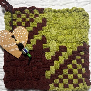 Op de afbeelding: Een vierkante geweven pannenlap met een geometrisch patroon in donker bordeauxrood en limoengroen. Een hartvormig kartonnen label is bevestigd met een zwart touwtje, met de tekst "Handmade with love" en drie decoratieve kralen van hout, zwart en gestreept glas.