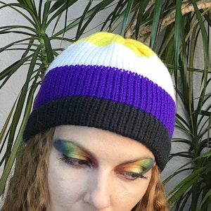 Peut inclure: Un bonnet en tricot à rayures blanches, jaunes, violettes et noires. Le bonnet est porté par une personne aux cheveux longs, bouclés, brun rougeâtre et au maquillage des yeux coloré.