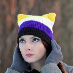 May include: A person wearing a gray coat with a purple, yellow, and black striped knit hat with cat ears.