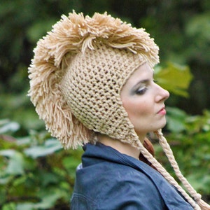 May include: A woman wearing a beige crocheted hat with a large, spiky mohawk. The hat has long, dangling ties.