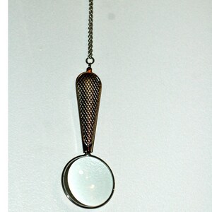 May include: A silver-toned magnifying glass pendant suspended from a delicate metal chain. The handle features a textured, cross-hatched diamond pattern, while the circular lens is framed in a smooth, polished silver-colored metal rim.