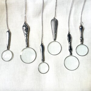May include: Six silver-toned magnifying glass pendants hanging on thin metal chains. Each pendant features a unique handle design, ranging from smooth and plain to ornate floral patterns or cross-hatched textures, attached to a circular lens.
