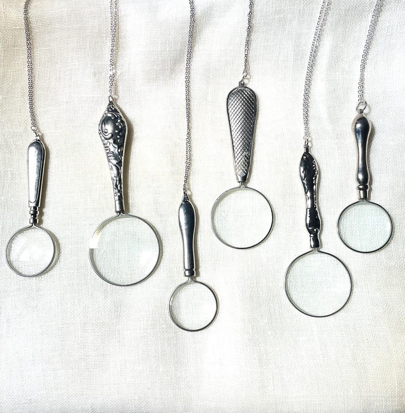 May include: Six silver-toned magnifying glass pendants hanging on thin metal chains. Each pendant features a unique handle design, ranging from smooth and plain to ornate floral patterns or cross-hatched textures, attached to a circular lens.