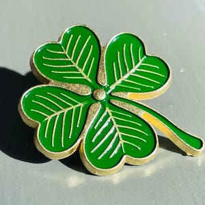 Puede incluir: Un pin de trébol de cuatro hojas de color dorado con esmalte verde. El trébol tiene un diseño detallado con líneas en cada hoja.