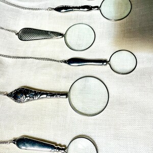 May include: Six silver-toned magnifying glass pendants on thin metal chains. Each pendant features a circular lens with a distinct handle design, including smooth, textured, and ornate patterns, arranged vertically on a light-colored fabric surface.