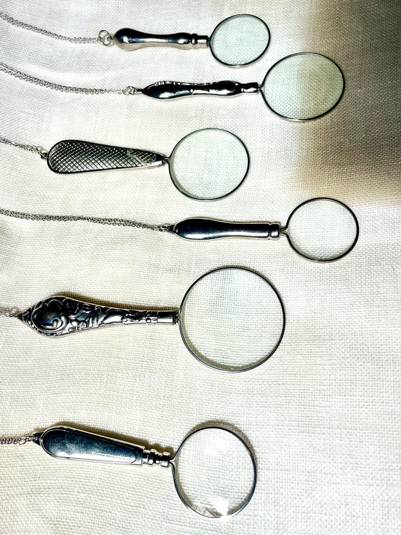 May include: Six silver-toned magnifying glass pendants on thin metal chains. Each pendant features a circular lens with a distinct handle design, including smooth, textured, and ornate patterns, arranged vertically on a light-colored fabric surface.