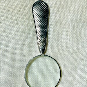 May include: A silver-toned magnifying glass pendant hanging from a thin metal chain. The handle features a textured, cross-hatched diamond pattern, while the circular lens is framed in a smooth, polished silver-colored metal rim.