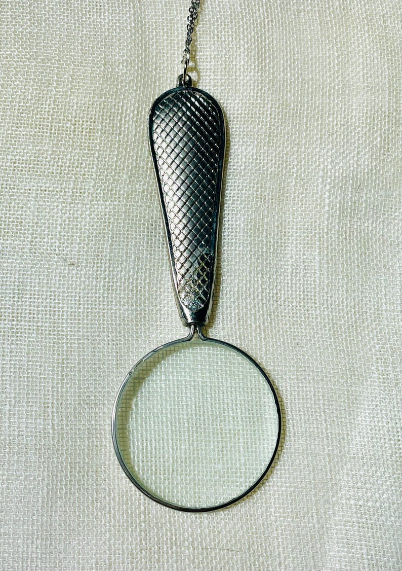May include: A silver-toned magnifying glass pendant hanging from a thin metal chain. The handle features a textured, cross-hatched diamond pattern, while the circular lens is framed in a smooth, polished silver-colored metal rim.